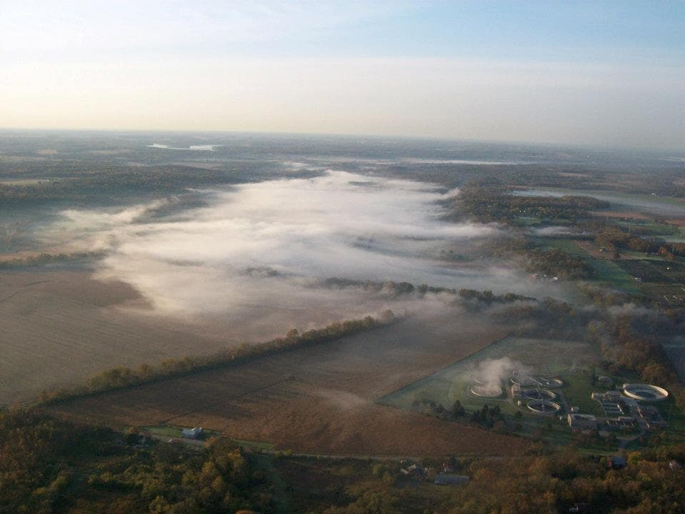 Aerial view of thick white fog settling over rural fields and forests at dawn.