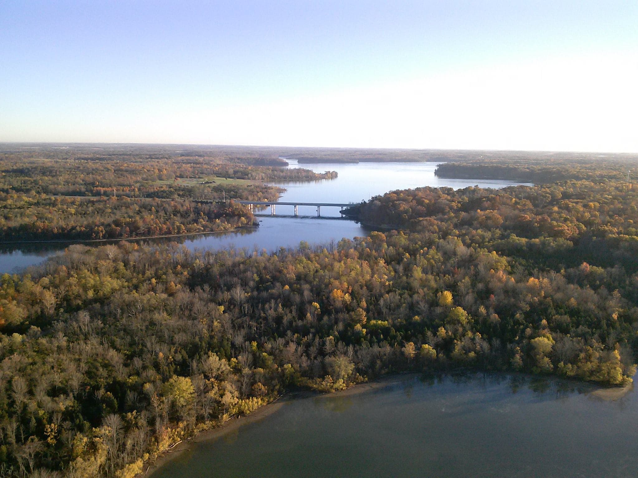 Aerial view of a bridge spanning a lake surrounded by dense, colorful autumn forests.