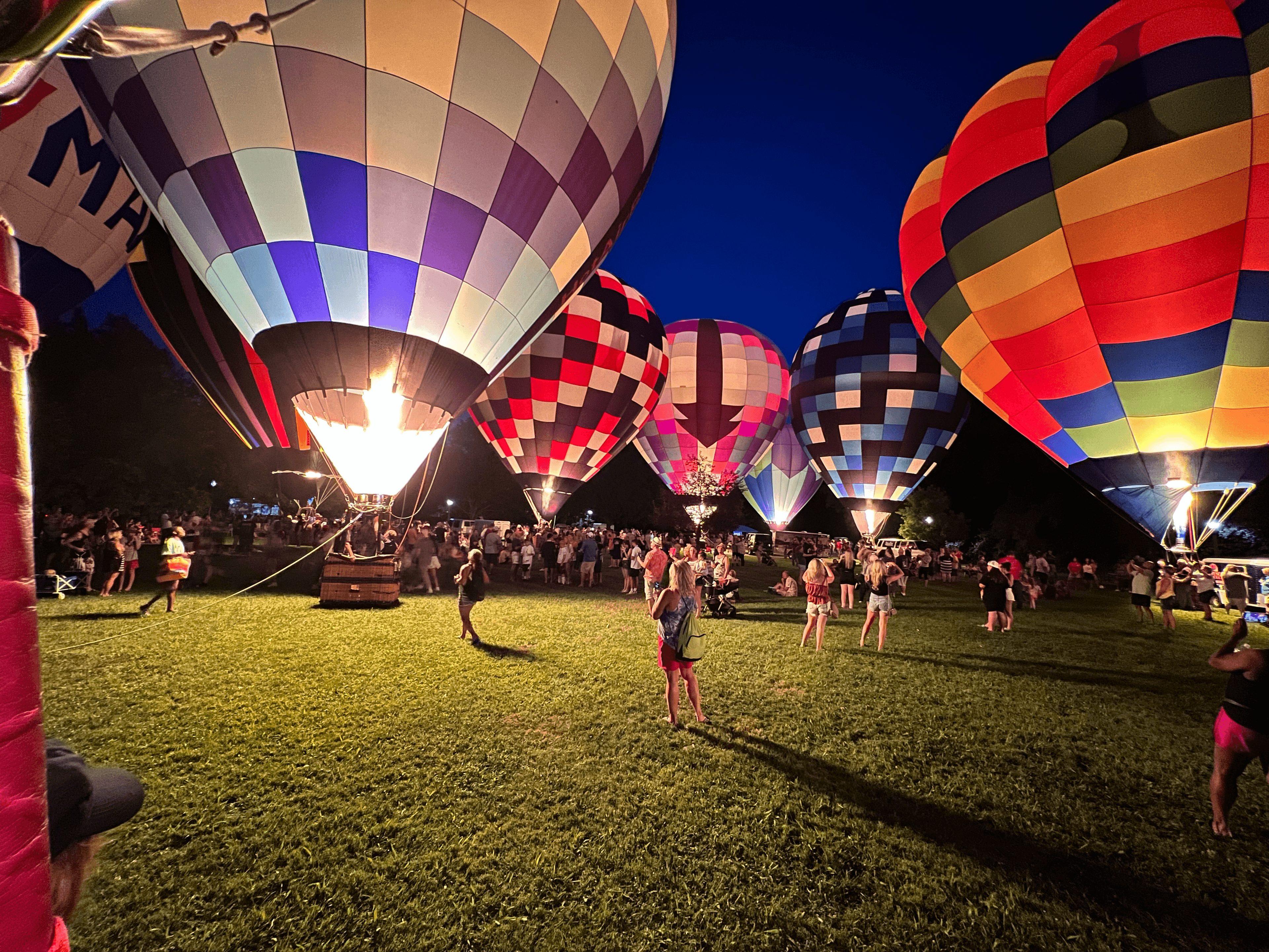 Vibrant hot air balloons glow at night over a grassy field filled with spectators.